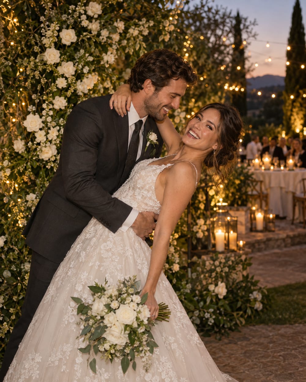 Bride and groom posing in front of a white rose and greenery backdrop at dusk